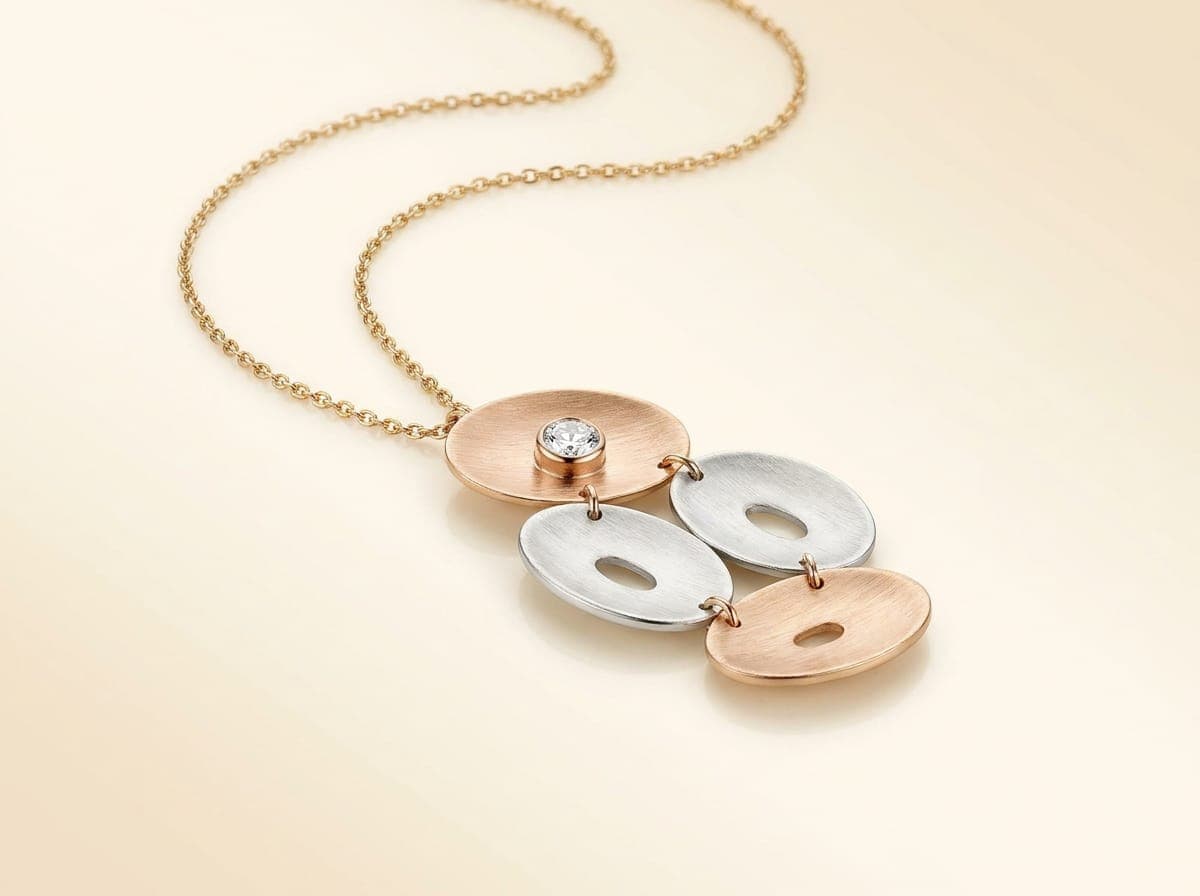 YUG pendant on a fine gold chain: overlapping brushed oval discs in rose gold and white gold with a single round Swarovski Zirconia on the top disc, on an ivory counter